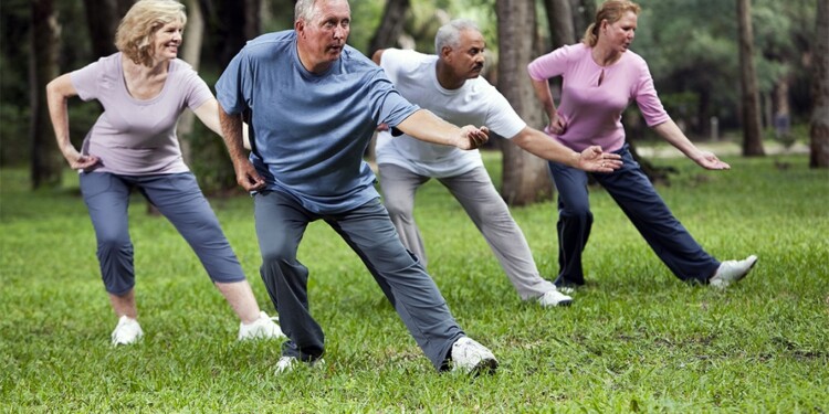 Multi-ethnic group of adults practicing tai chi in park.  Main focus on senior man (60s) in blue shirt.