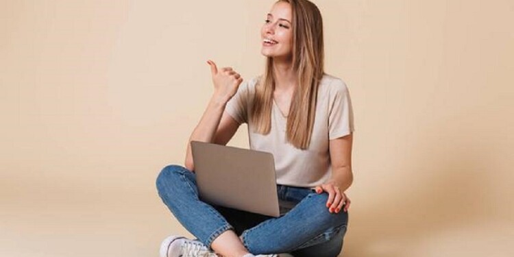 Portrait of a smiling casual girl with laptop computer sitting on a floor with legs crossed and pointing finger away isolated over beige background