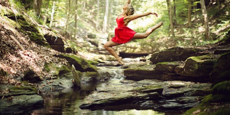 Beautiful girl ballerina, dancing ballet in green forest next the creek. She is jumping over the stream and she spread hers arms as flying. Girl is wearing a short red dress, has long blond hair and she is barefoot. She is smiling and she is hapy while dancing.
