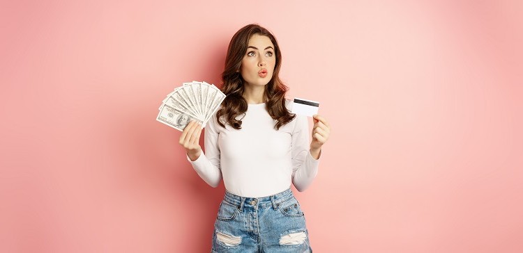 Credit, money and loans concept. Happy beautiful girl holding credit card and cash, looking satisfied, thinking of shopping, standing over pink background.