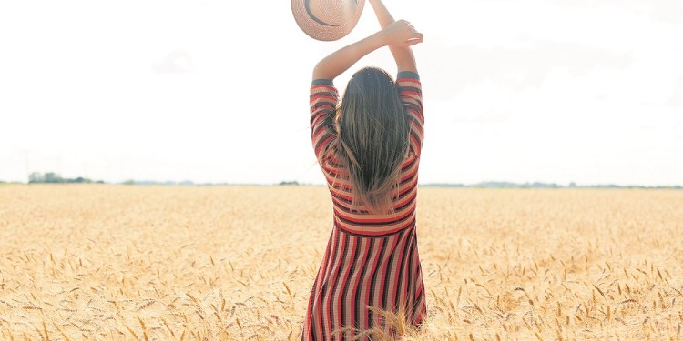 Backview of slender well shaped woman holding her arms up, having straw hat in one hand, standing in front of Sun with pleasure in middle of wheat field, enjoying summer holidays at rural area.