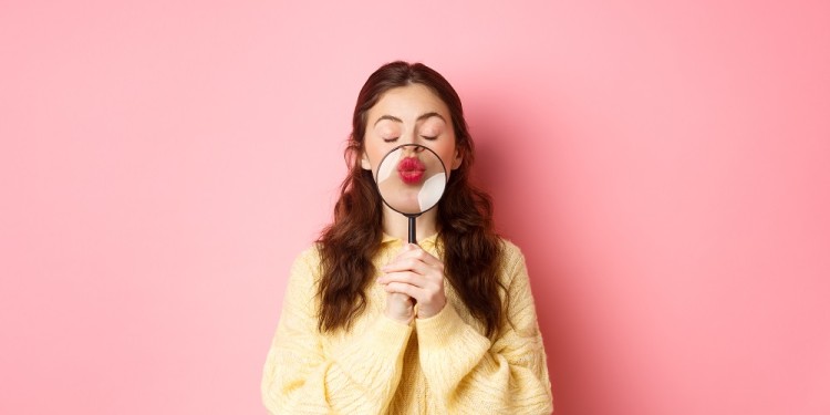 Cute and romantic young woman magnify her lips with magnifying glass, making kissing face, standing with closed eyes against pink background.