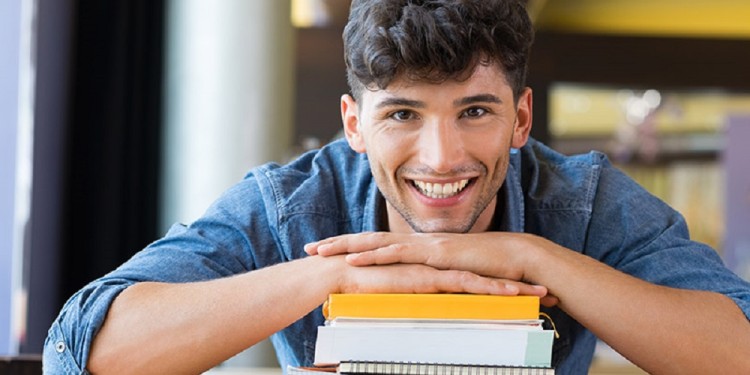 Closeup shot of young man leans on stack of books. Happy male student smiling and looking at camera. Lughing guy leaning on a pile of books and school notebooks.
